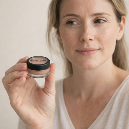 Woman holding a jar of Omiana concealer cream against a plain background